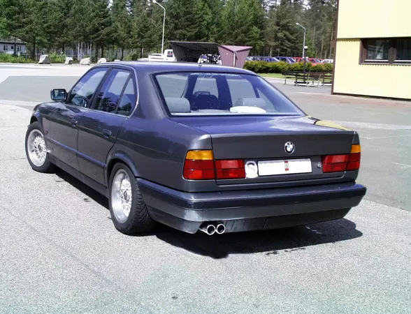 Vista posteriore di una BMW E34 grigio scuro. Si può notare il sistema di scarico sportivo duplex in acciaio cromato con terminali da 2x70 mm, installato sotto il paraurti. Il veicolo è parcheggiato in un parcheggio, con alberi e un edificio sullo sfondo.