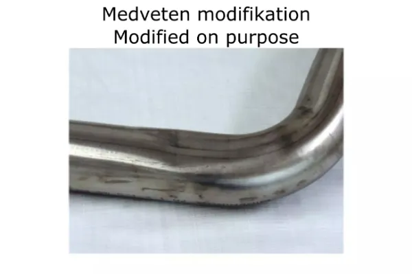 Primo piano di un tubo in acciaio inox piegato per scarico sportivo Simons. Argento lucido con lievi segni di usura. Sul tubo: Modifica Medveten Modificato di proposito. Componente per BMW F30 320i, 328i.